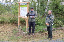 Zwei Männer in Dienstkleidung stehen im Wald vor einem Schild