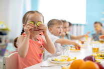 Mädchen an Essenstisch mit mehreren Kindern hält sich Gurkenscheiben vor Augen. © Oksana Kuzmina - stock.adobe.com