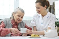 Frau reicht einer Seniorin Tasse auf Untertasse, davor auf dem Tisch Kuchenstück und Kaffeekanne